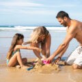 Joyful mom, dad and little daughter enjoying vacation at sea together, playing with daughters sand toys, building sandcastle. Family summer holidays concept