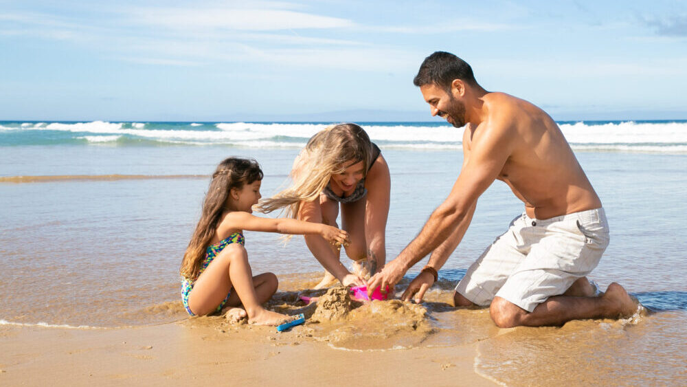 Joyful mom, dad and little daughter enjoying vacation at sea together, playing with daughters sand toys, building sandcastle. Family summer holidays concept